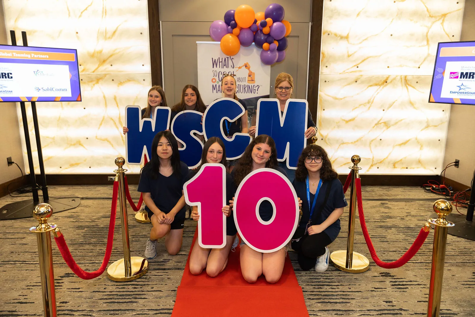 Students kneeling on a red carpet behind giant WSCM and 10 letters between gold stanchions and velvet ropes