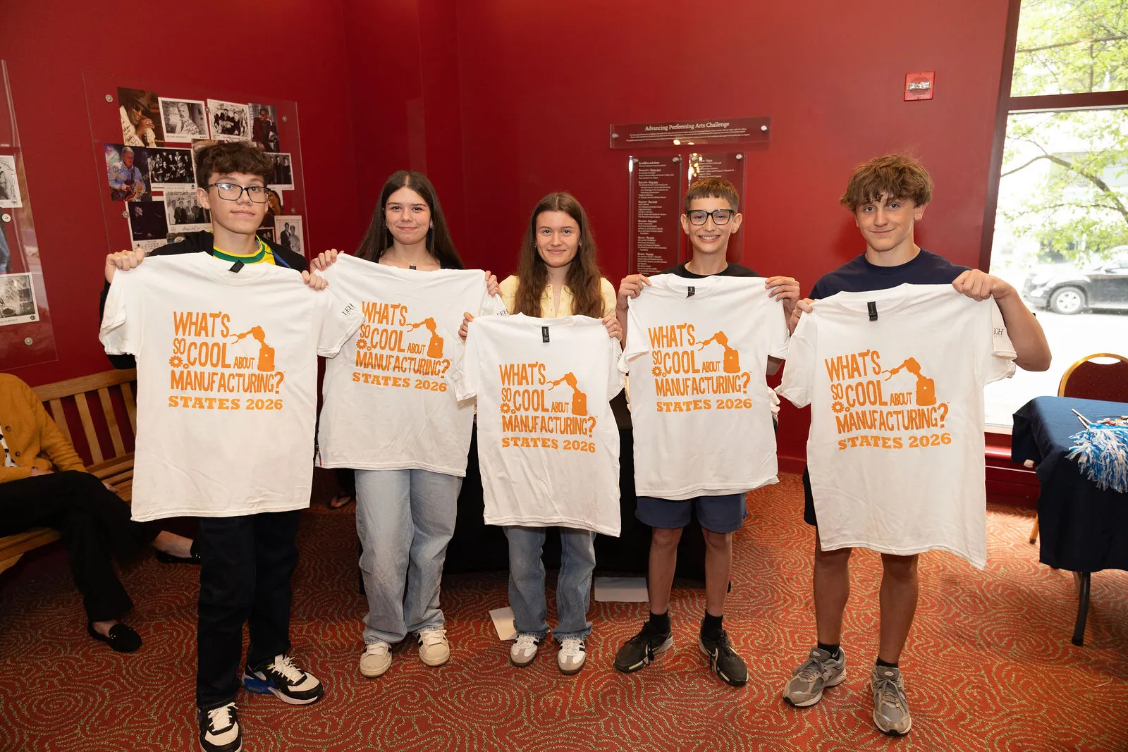 Students holding up What's So Cool About Manufacturing States 2026 T-shirts in the lobby