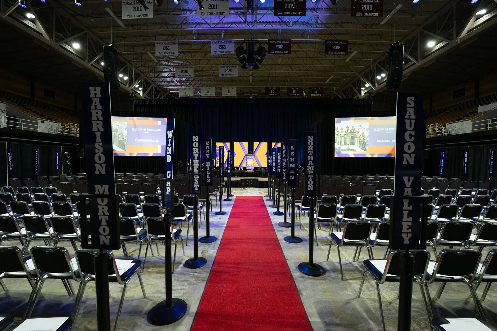 The empty arena floor with rows of chairs, a red carpet down the center aisle and school banners hanging over the rows